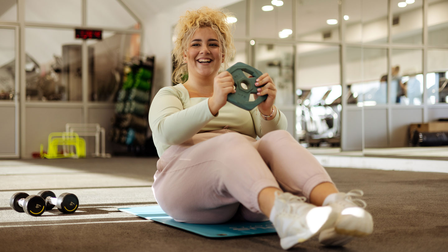 Mujer sonriente en un gimnasio sosteniendo una pesa, usada como ilustración de una nota sobre preservación de masa magra durante el adelgazamiento