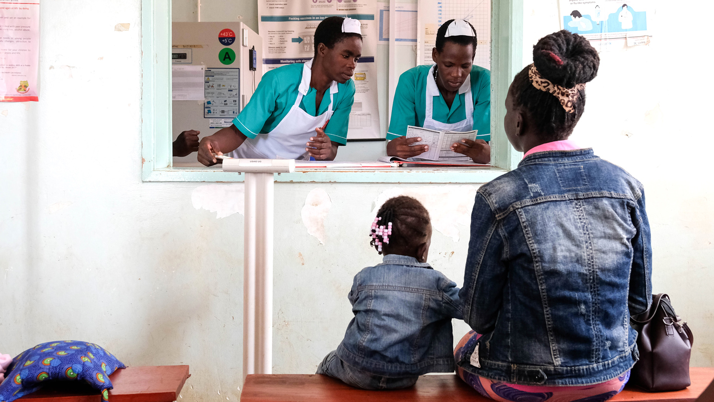 Una madre y una niña esperan atención mientras personal sanitario revisa documentos en un centro de salud de Uganda.